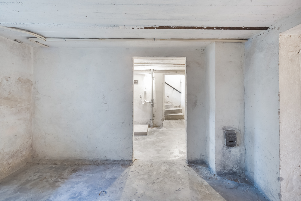 Leerer, heller Kellerraum mit sichtbaren Rohrleitungen und einer Tür zu einer Treppe.