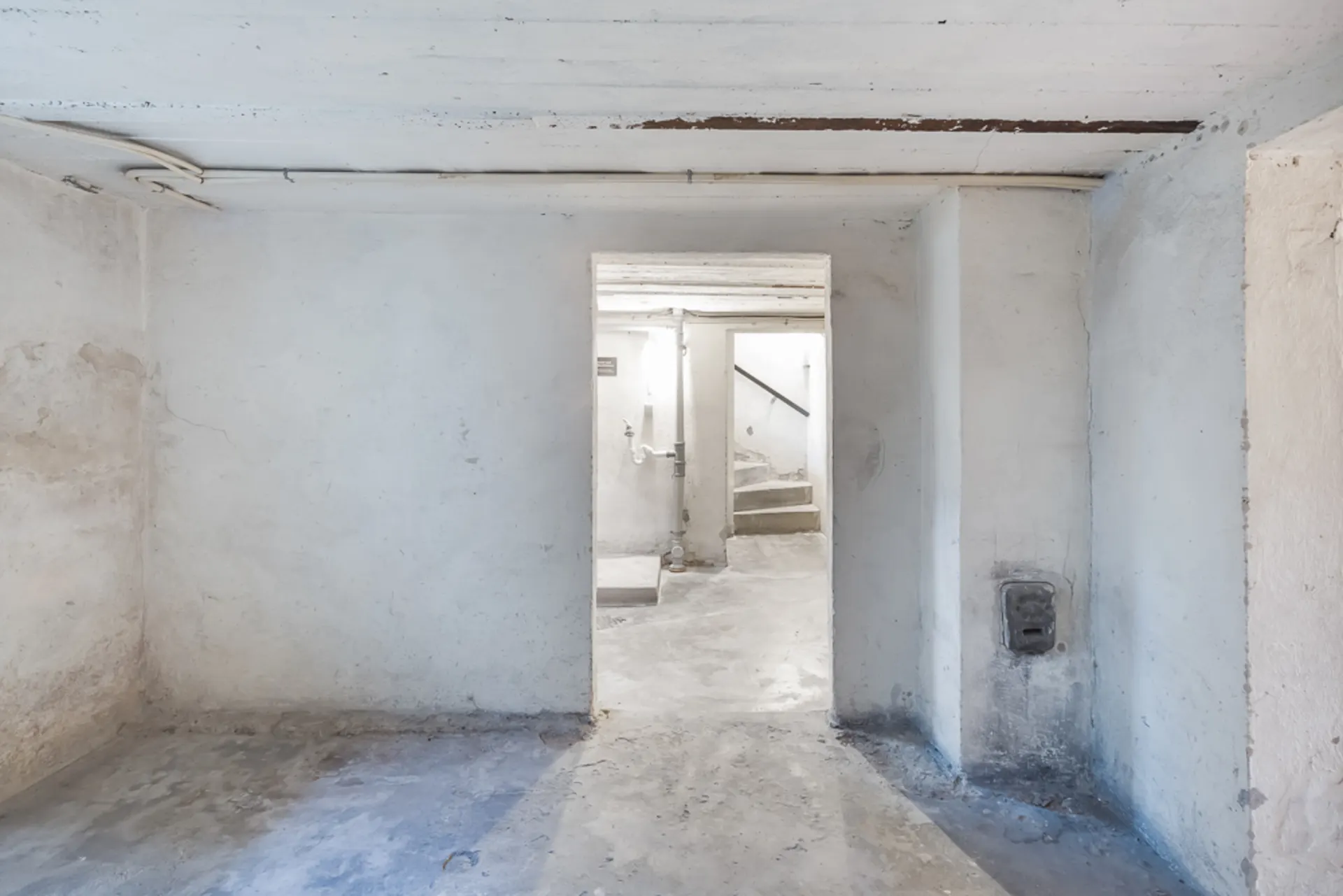 Leerer, heller Kellerraum mit sichtbaren Rohrleitungen und einer Tür zu einer Treppe.