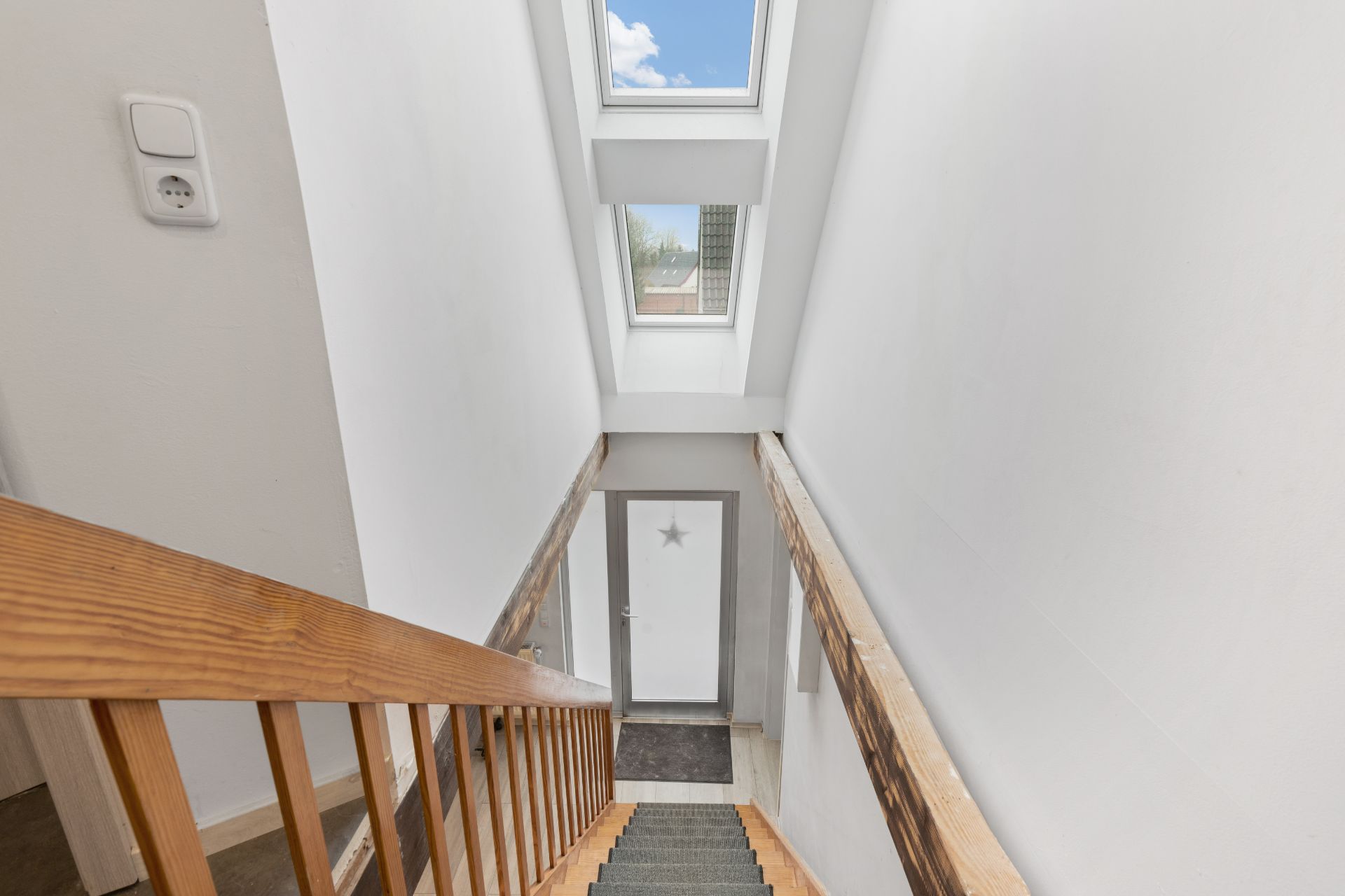 Treppe mit Holzgeländer, Oberlichtfenster und weißer Wand in einem Hausflur.