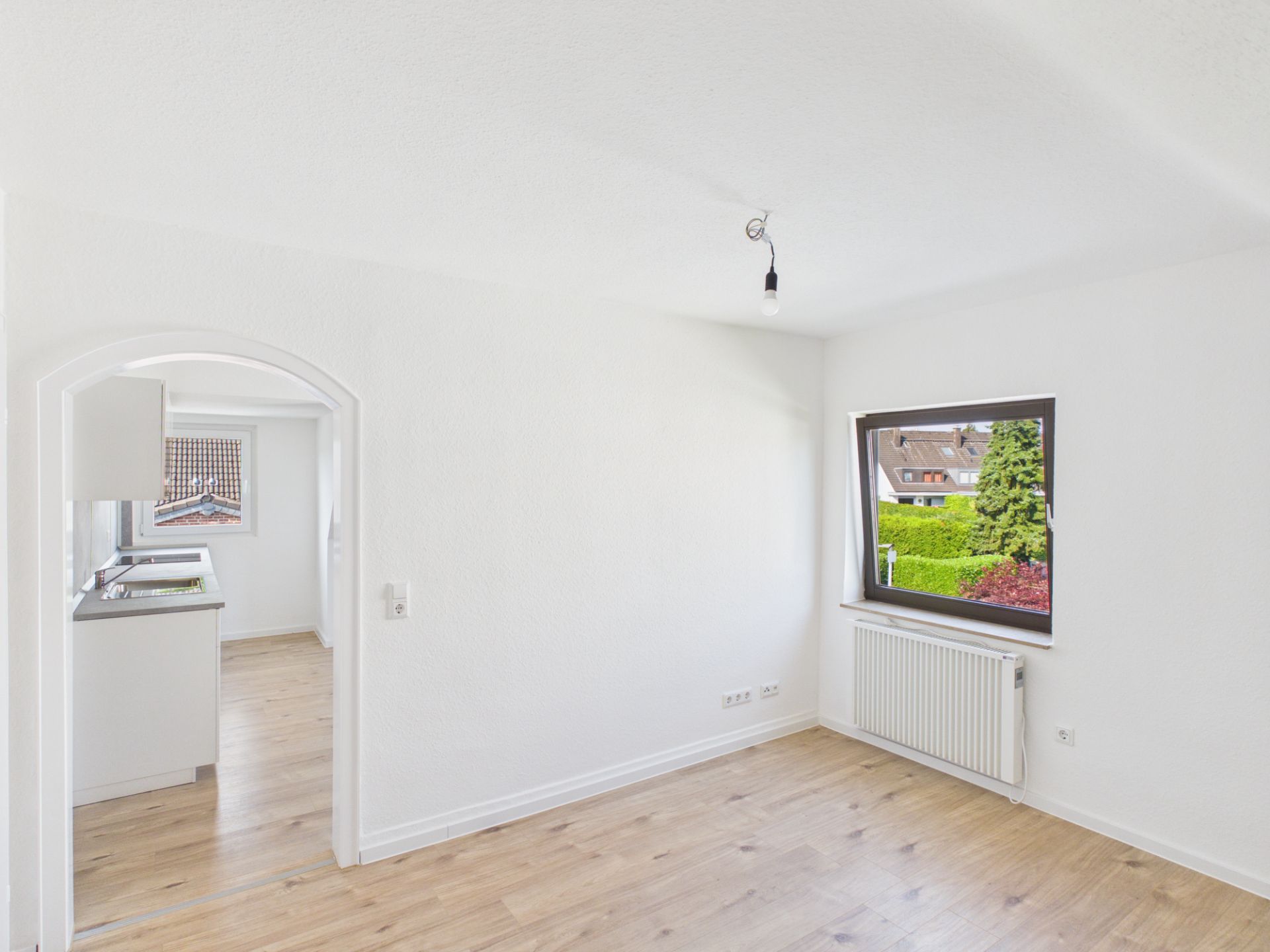 Helles Zimmer mit Holzfußboden, Fenster und Zugang zur Küche durch einen Rundbogen.