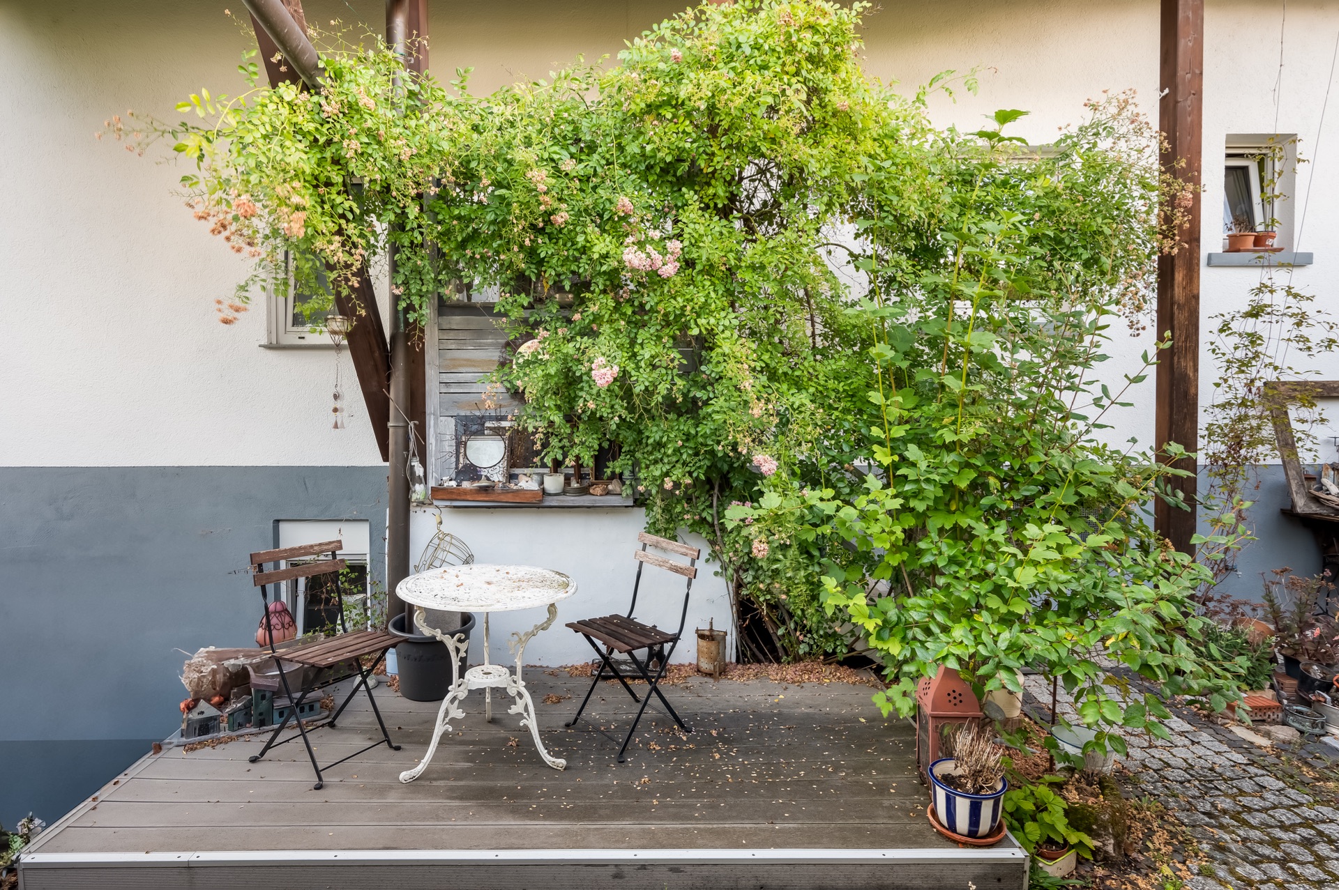 Kleine Terrasse mit zwei Stühlen, rundem Tisch und viel Grünpflanzen an einer Hauswand.