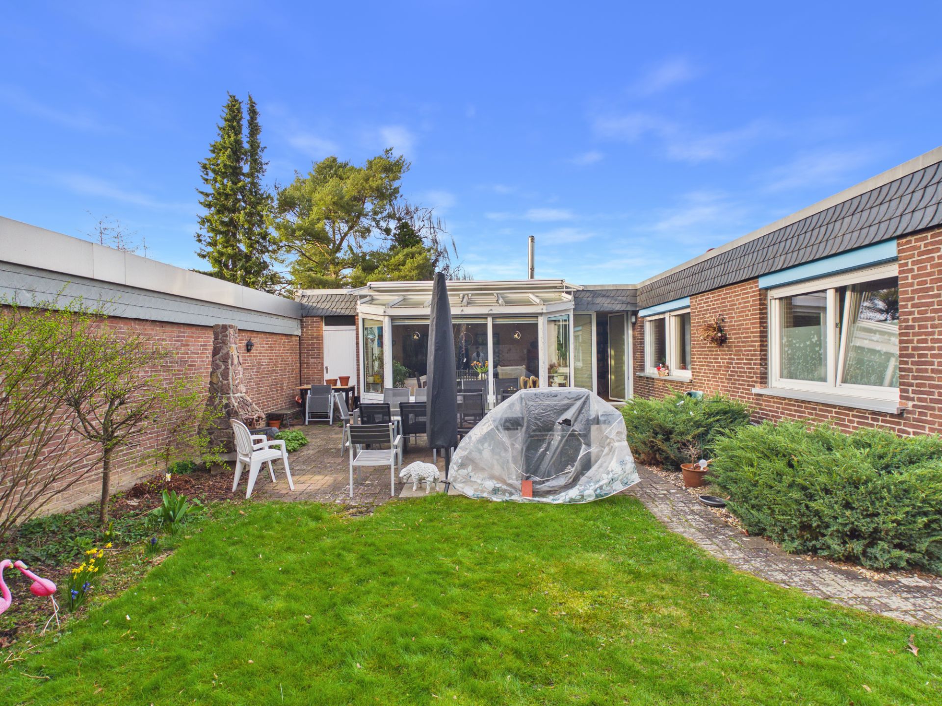 Gepflegter Garten mit Terrasse, Gartenmöbeln und Ziegelhaus im Hintergrund.
