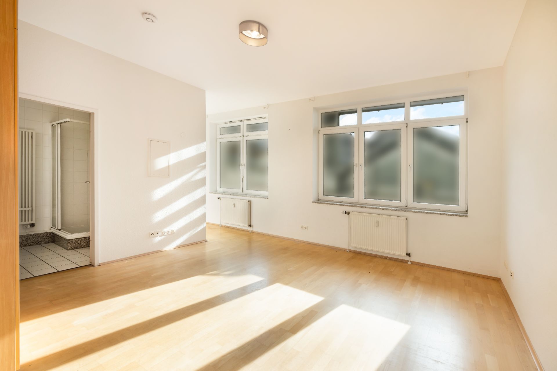 Helles, leeres Zimmer mit Holzboden, großen Fenstern und Zugang zu einem Badezimmer.