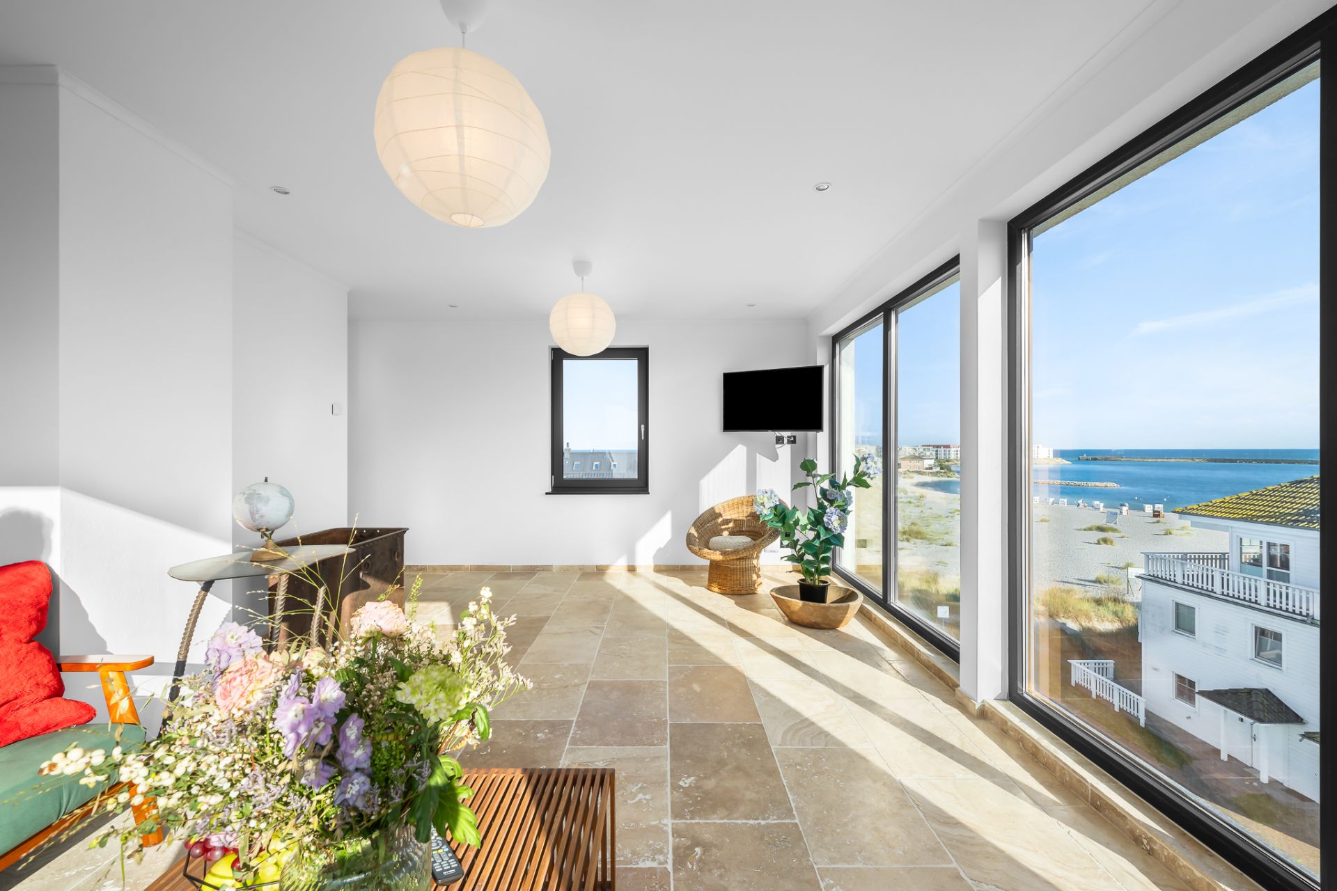 Helles Wohnzimmer mit großen Fenstern, Meerblick, Pflanzen und Sitzmöbeln.