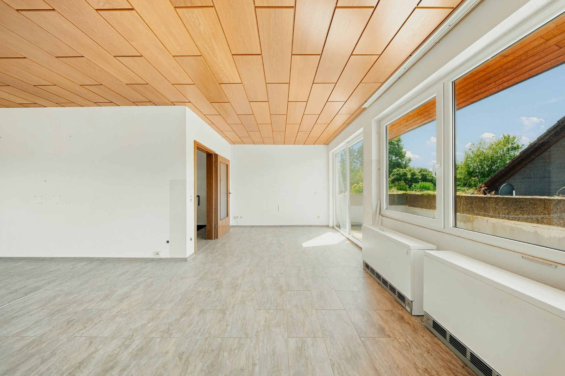 Heller, leerer Raum mit großen Fenstern, Holzdecke und Fliesenboden, Blick auf Garten.