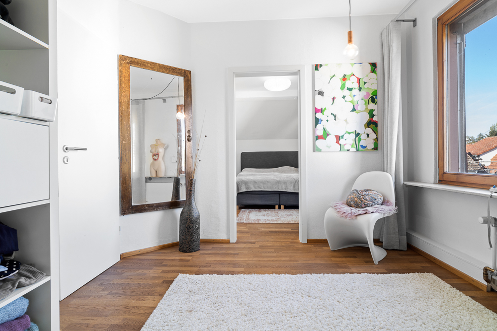 Heller Raum mit Spiegel, Stuhl, Teppich und Blick ins Schlafzimmer mit grauem Bett.
