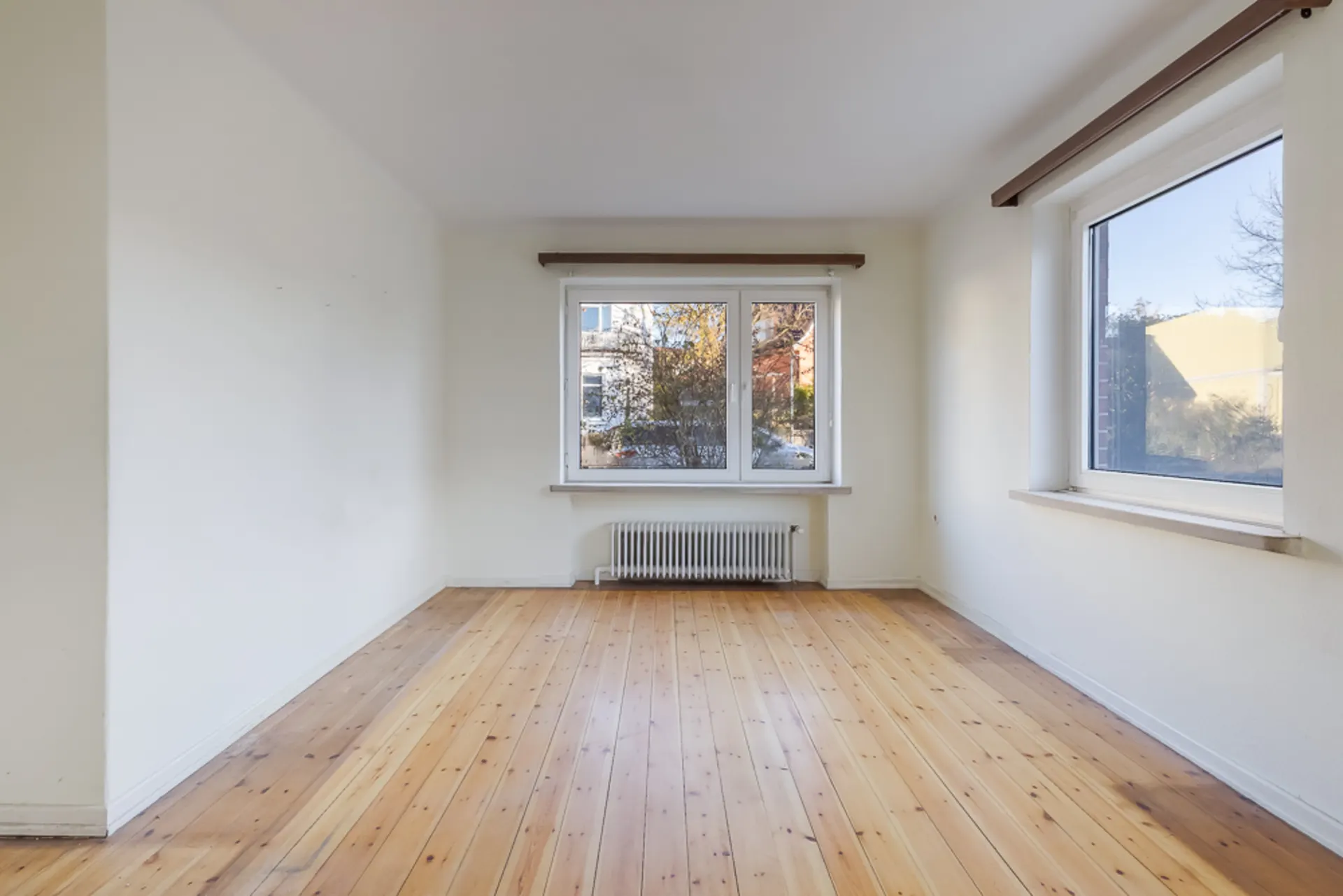 Leerer Raum mit Holzboden, zwei großen Fenstern und weiß gestrichenen Wänden.
