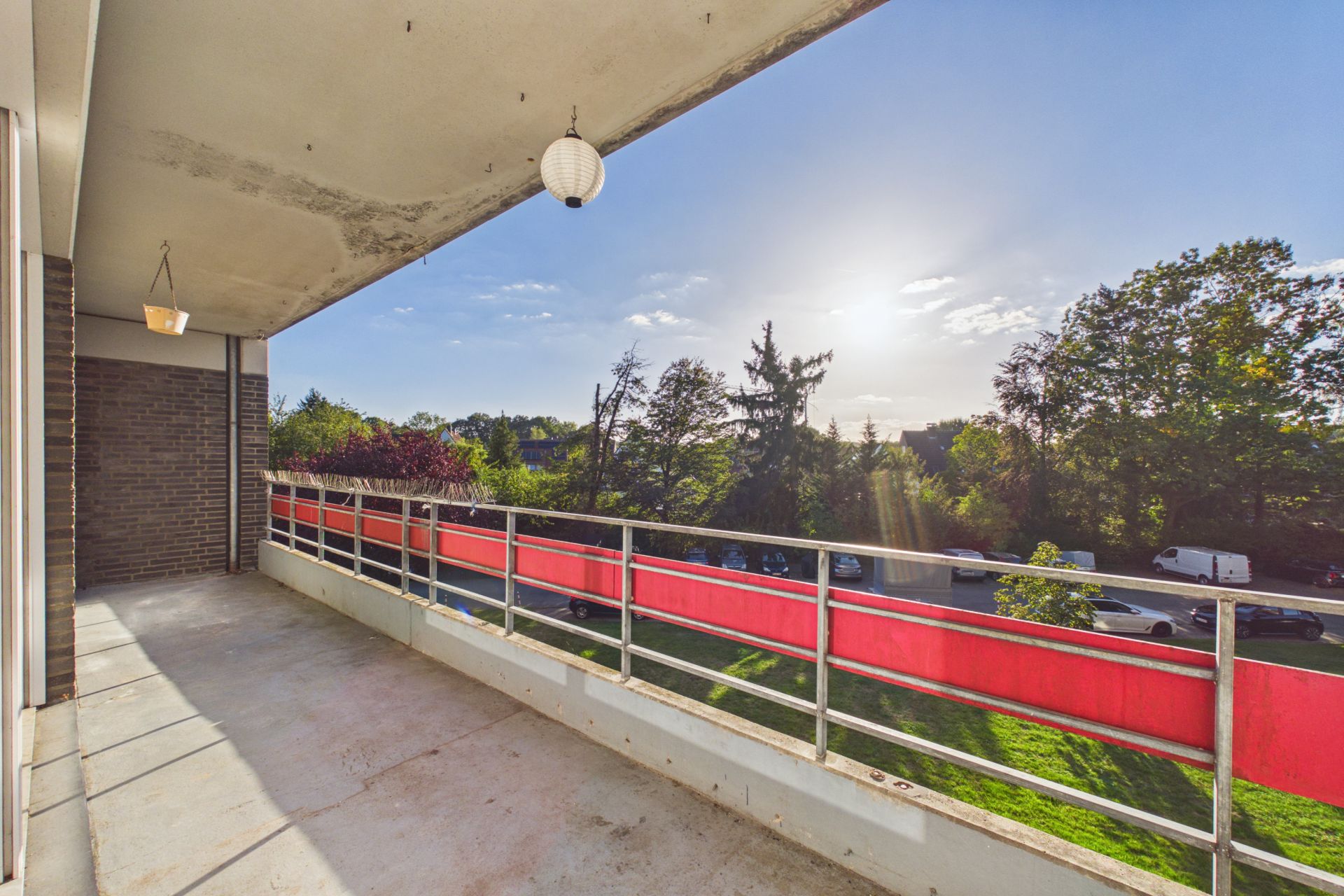 Großer Balkon mit rotem Geländer und Blick auf Bäume und Straße bei Sonnenschein.