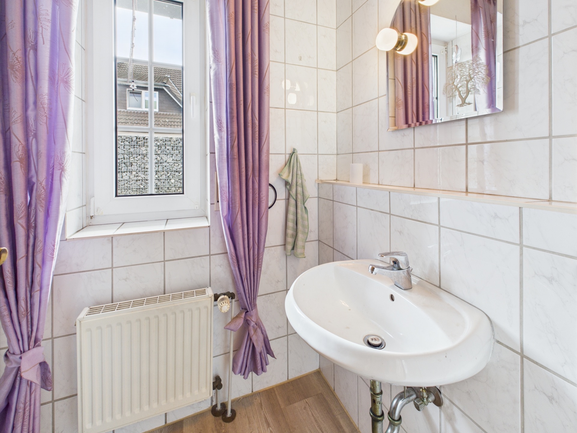 Helles Badezimmer mit Waschbecken, Fenster, lila Vorhängen und Wandspiegel.