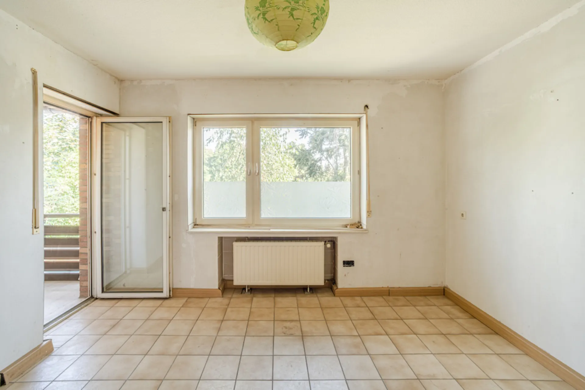 Leerer Raum mit Fliesenboden, großem Fenster, Heizung und offener Tür zum Balkon.