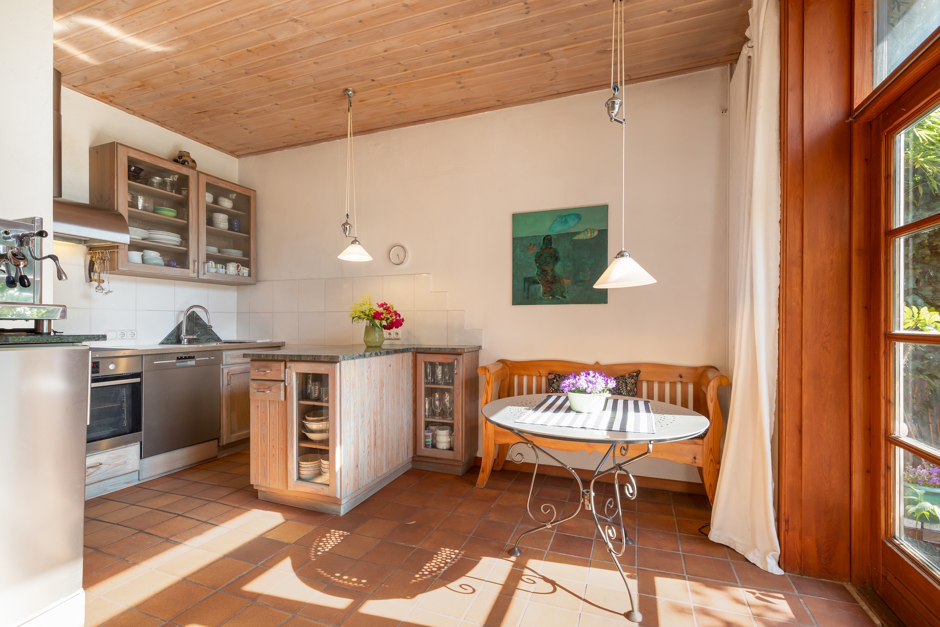 Helle Küche mit Holzmöbeln, rundem Tisch, Bank und großen Fenstern.
