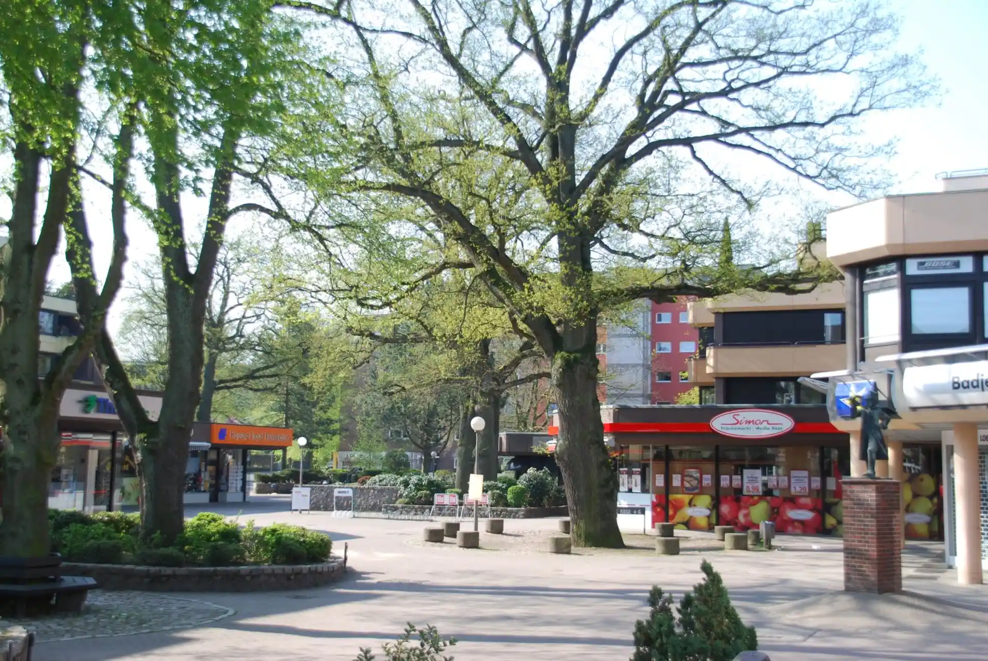 Platz mit Bäumen, Bänken und Geschäften, darunter ein Obstladen mit roter Markise.
