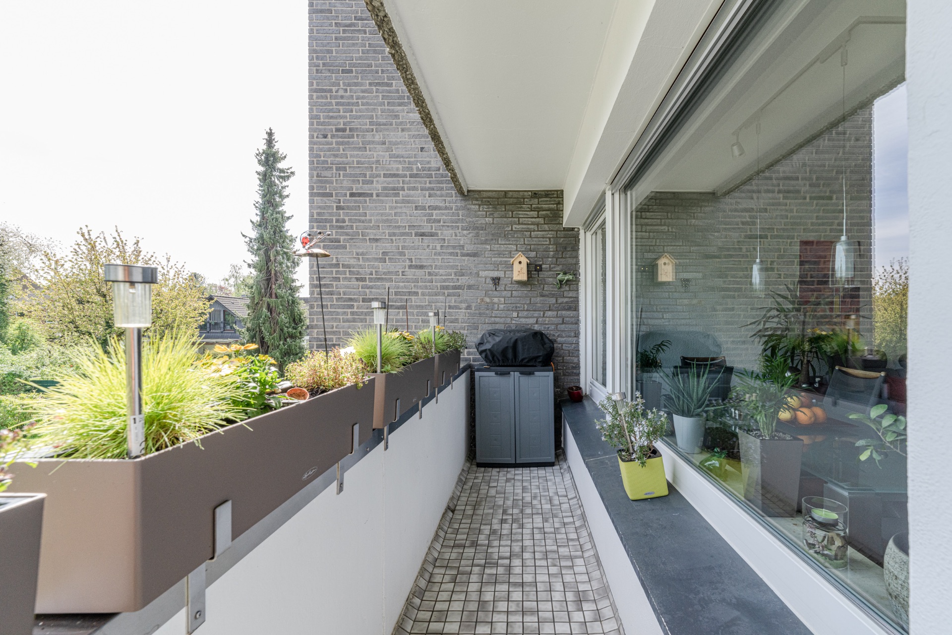 Schmaler Balkon mit Blumen, Topfpflanzen und einem Grill. Große Fensterfront zur Wohnung.
