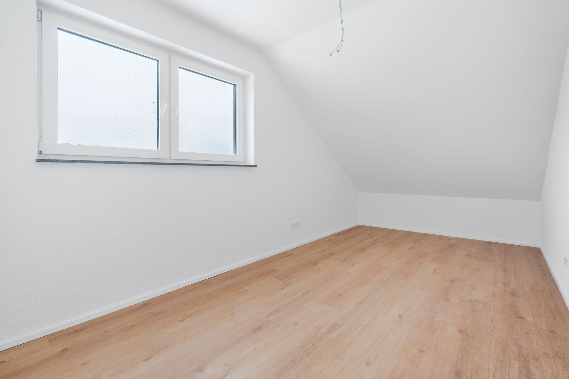 Leerer Raum mit schräger Decke, großem Fenster und hellem Holzfußboden.