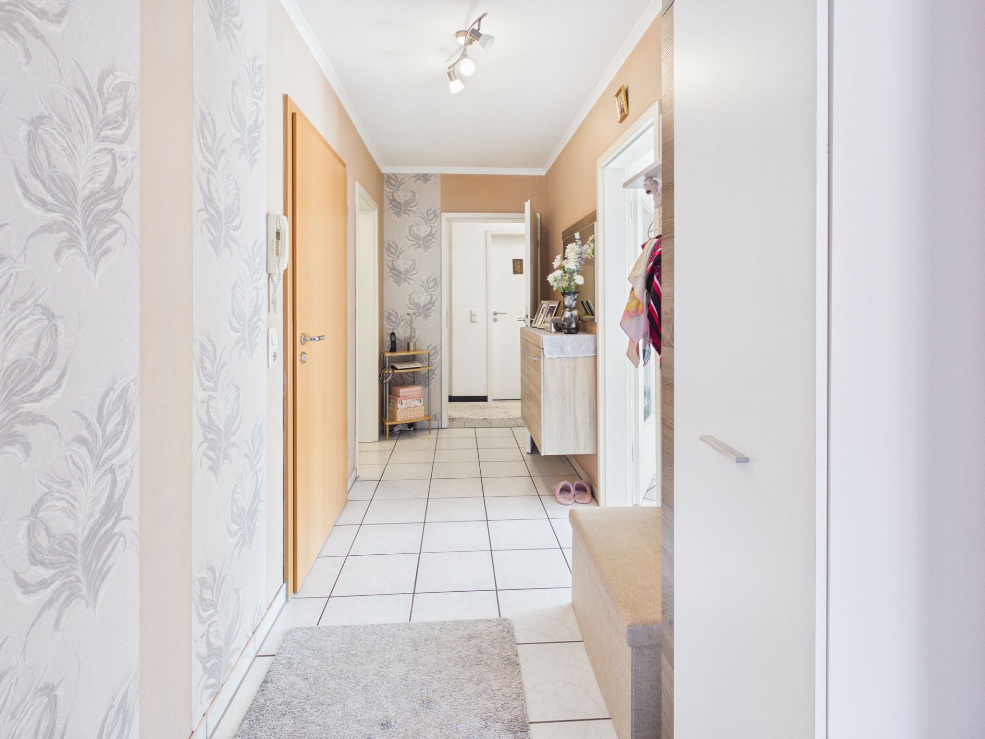 Heller Flur mit Fliesenboden, Garderobe, Kommode und Tapete mit Muster.