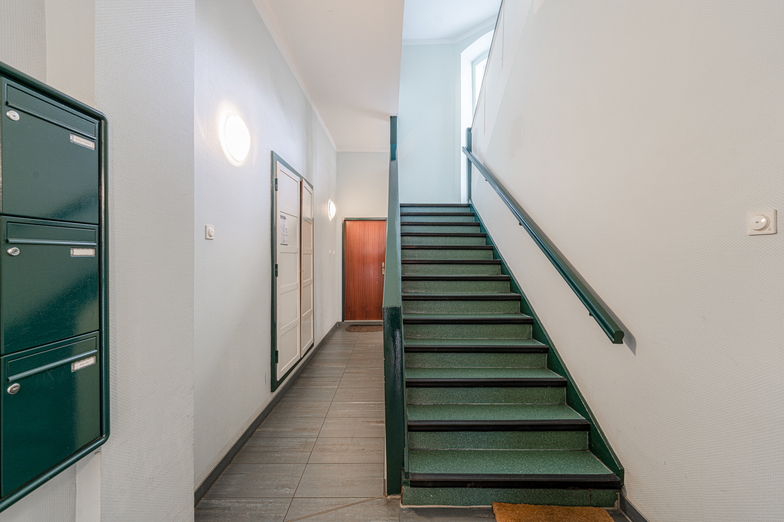 Hausflur mit grüner Treppe, Briefkästen und einer Holztür im Hintergrund.