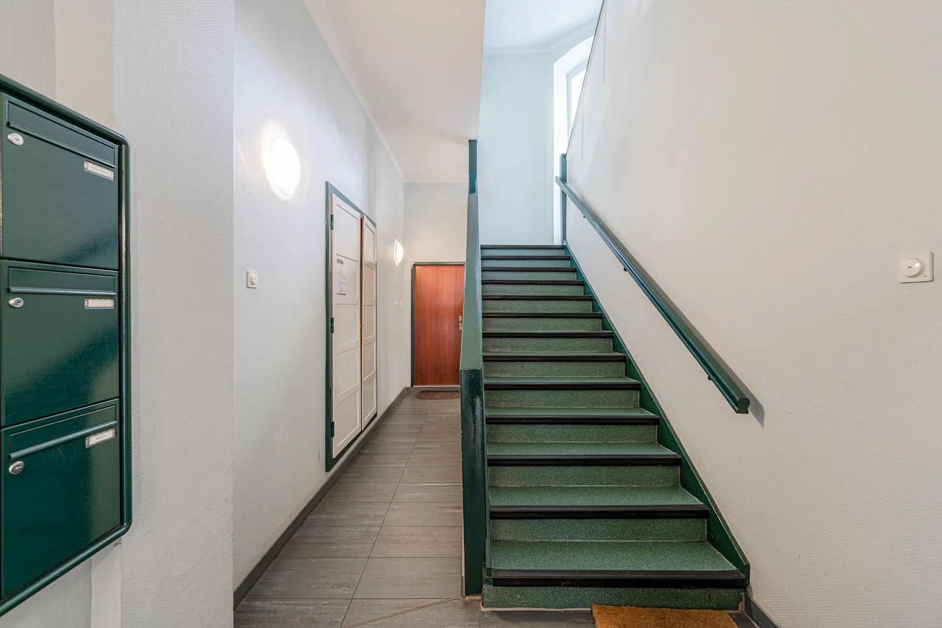 Hausflur mit grüner Treppe, Briefkästen und einer Holztür im Hintergrund.