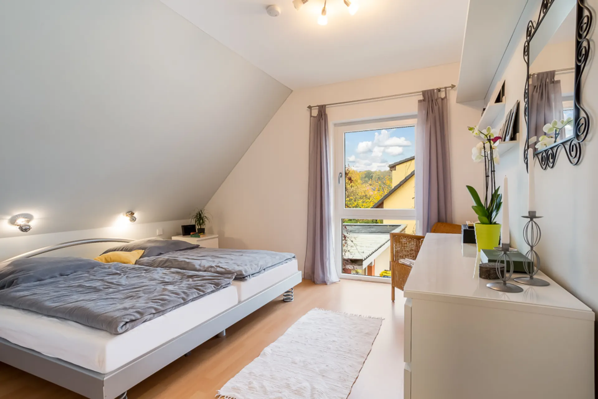 Helles Schlafzimmer mit Doppelbett, Kommode, Spiegel und Fenster mit Blick nach draußen.