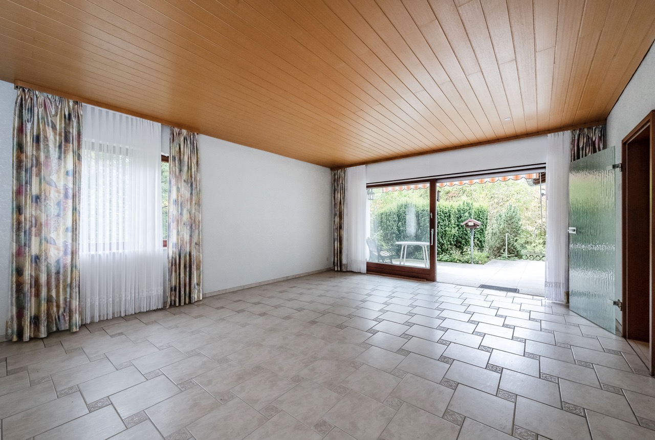 Leeres Wohnzimmer mit Fliesenboden, Holzdecke, großen Fenstern und Blick in den Garten.
