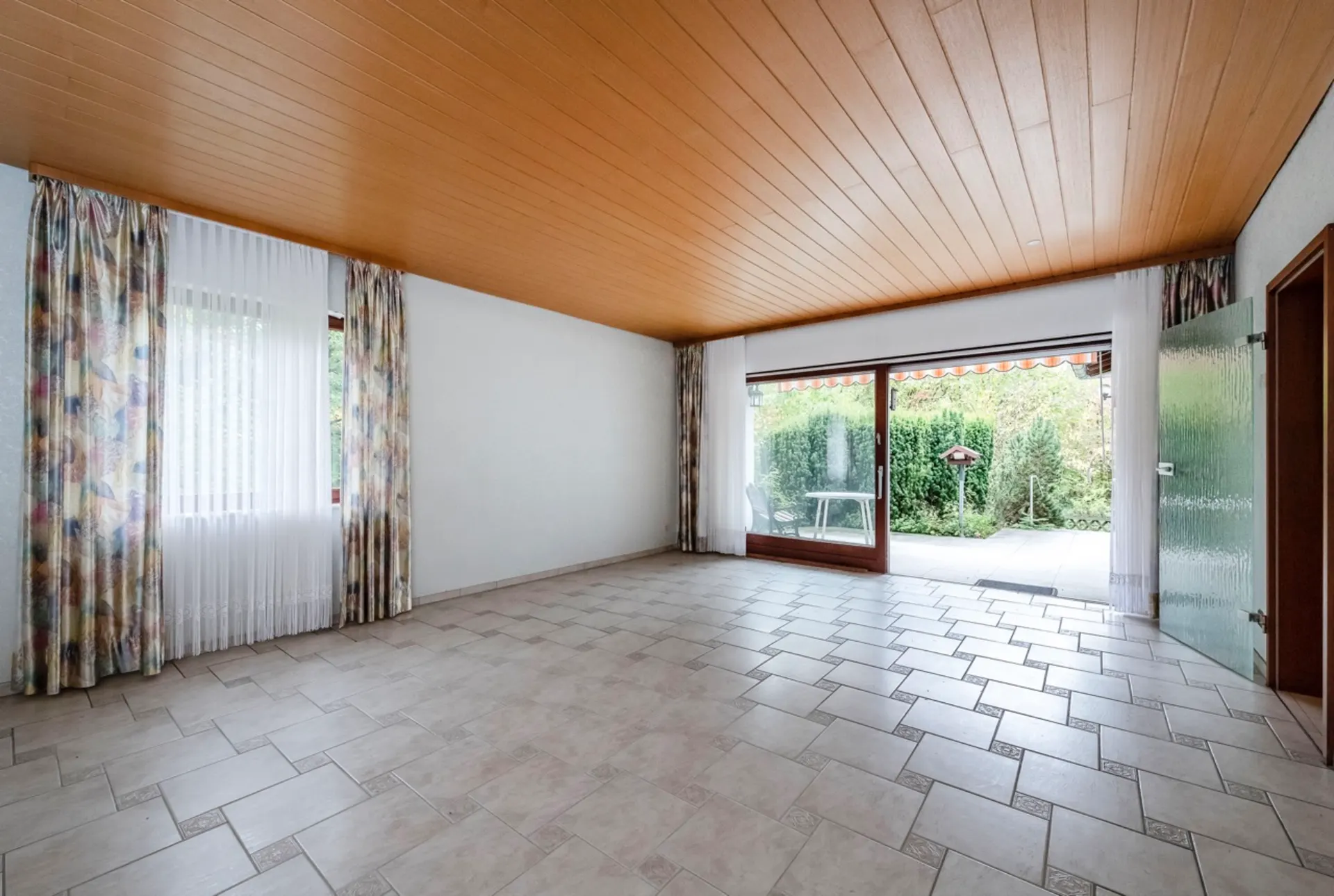 Leeres Wohnzimmer mit Fliesenboden, Holzdecke, großen Fenstern und Blick in den Garten.