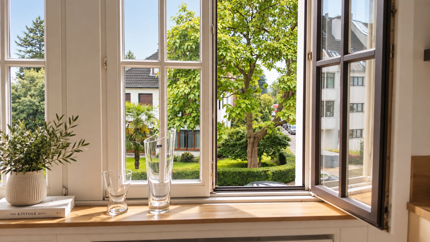 Offenes Fenster mit Blick auf Bäume und Wohngebäude, Vase und Gläser auf der Fensterbank.