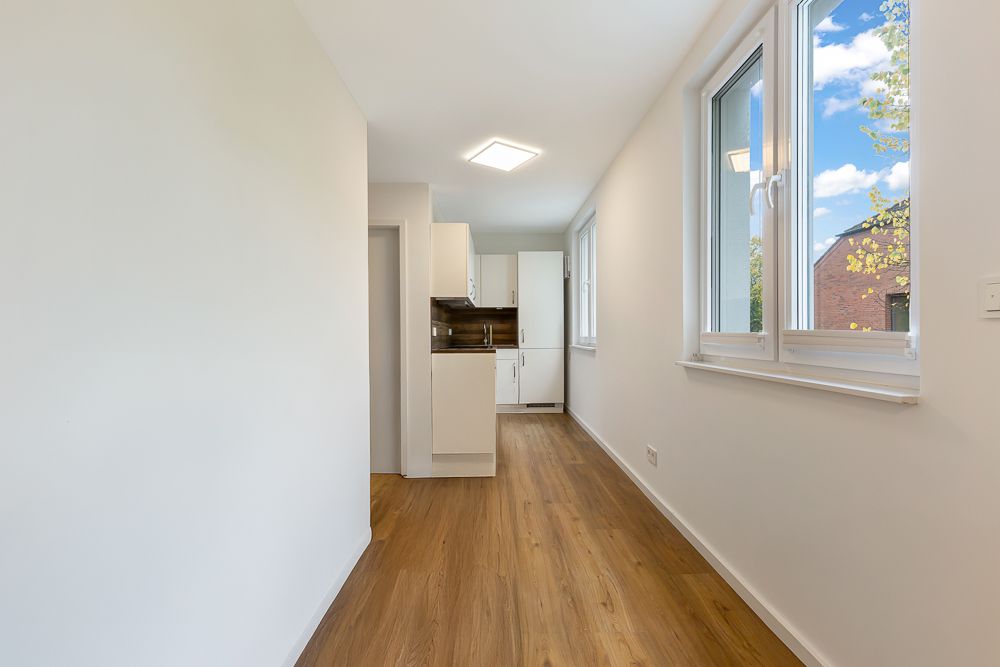 Heller Raum mit Holzboden, weißen Wänden, großen Fenstern und einer kleinen Einbauküche.