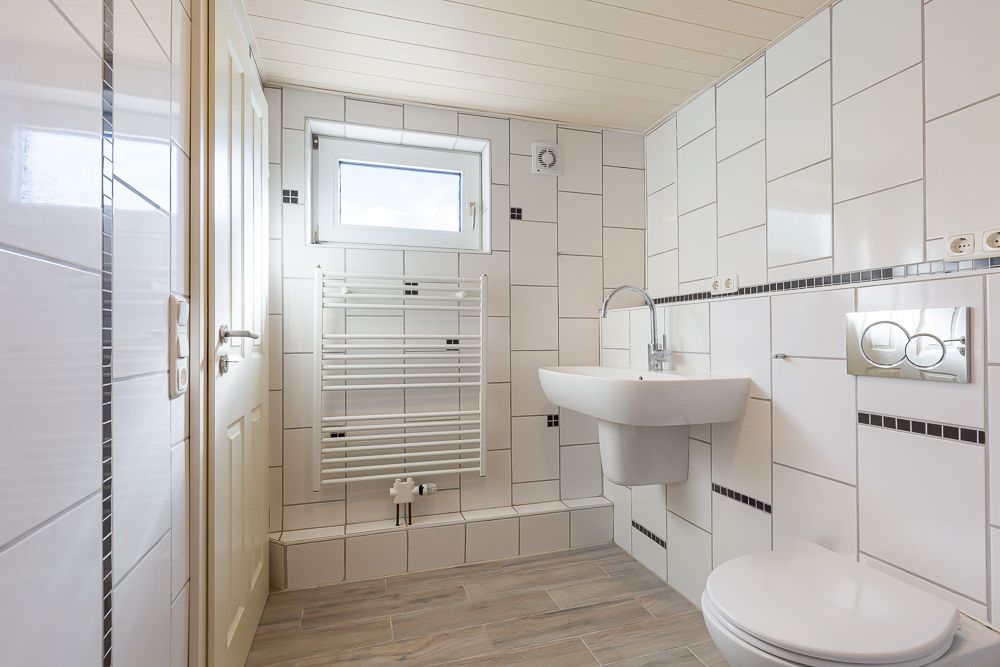 Helles, gefliestes Badezimmer mit Fenster, Waschbecken, Wandheizkörper und Toilette.
