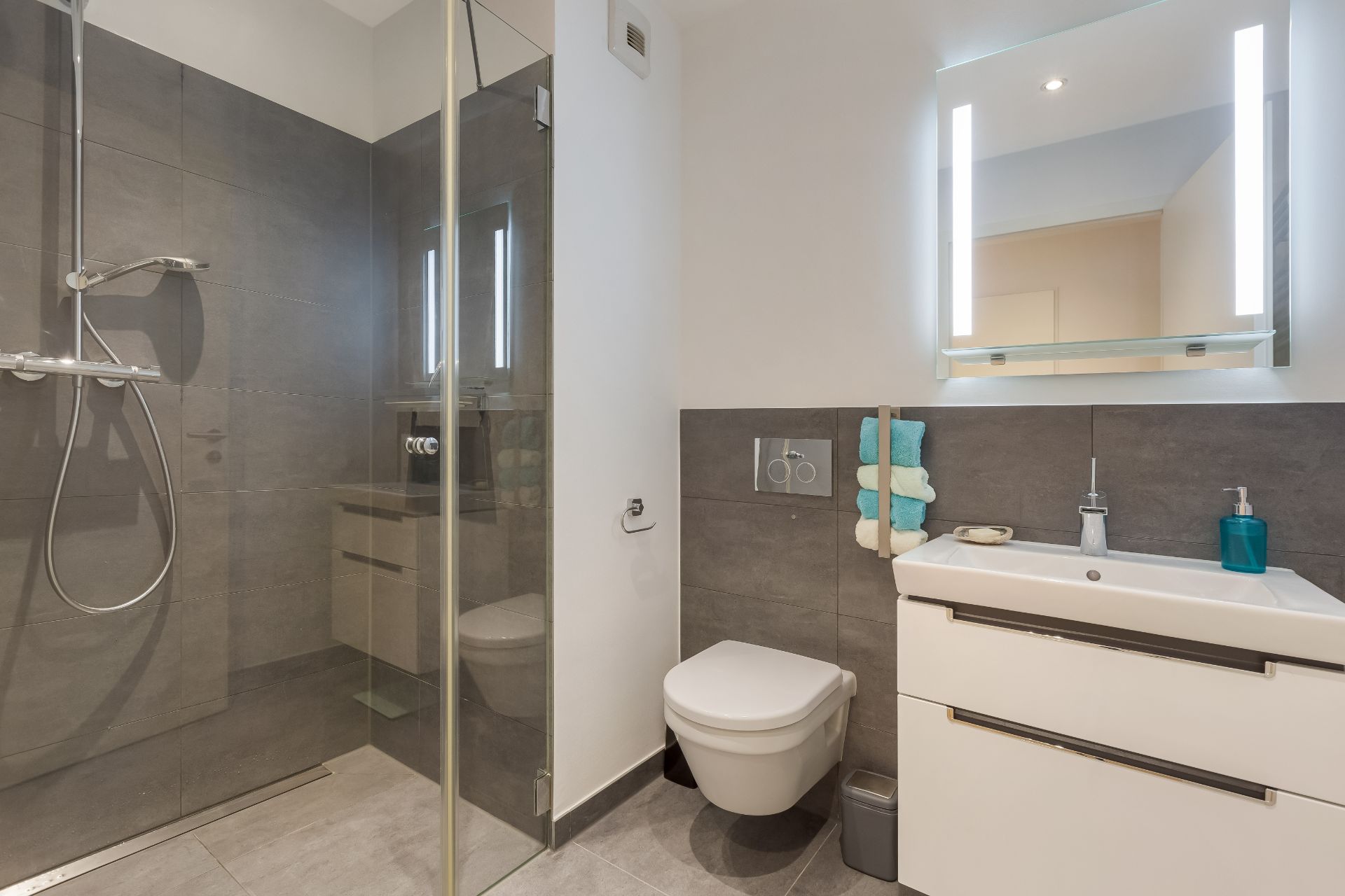 Modernes Badezimmer mit bodengleicher Dusche, WC, Waschbecken und beleuchtetem Spiegel.