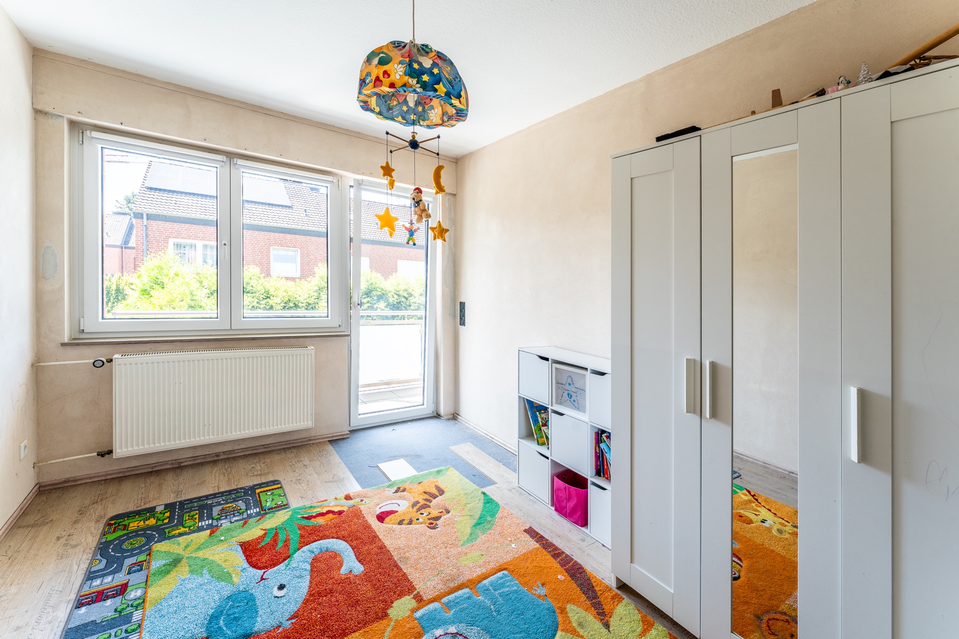 Helles Kinderzimmer mit buntem Teppich, weißem Schrank und großem Fenster.