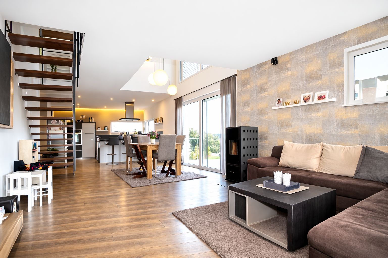 Modernes Wohnzimmer mit braunem Sofa, Couchtisch, Esstisch und offener Treppe, große Fenster zur Terrasse.