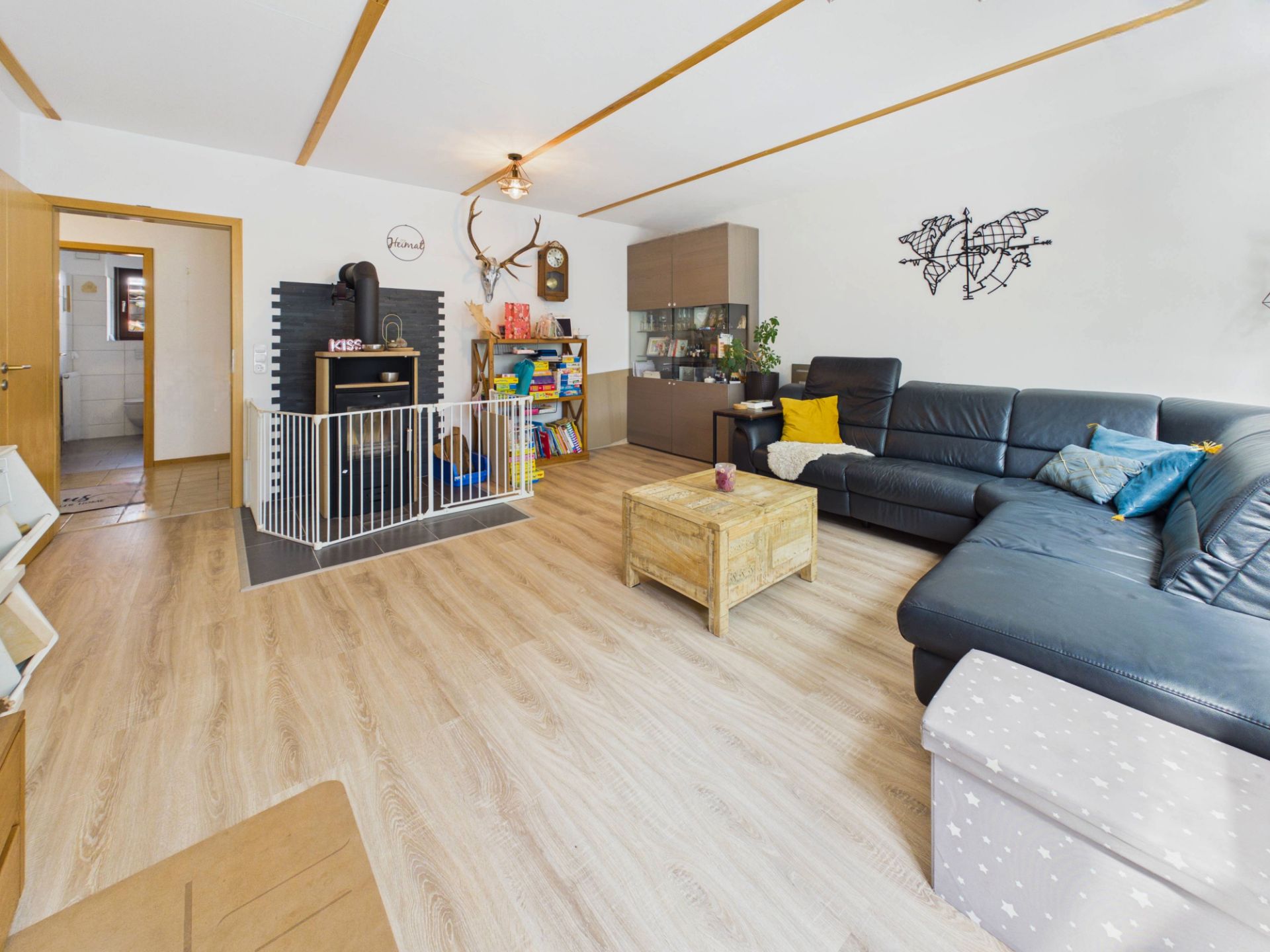 Helles Wohnzimmer mit großem Ecksofa, Holzofen mit Schutzgitter und Couchtisch auf Holzfußboden.