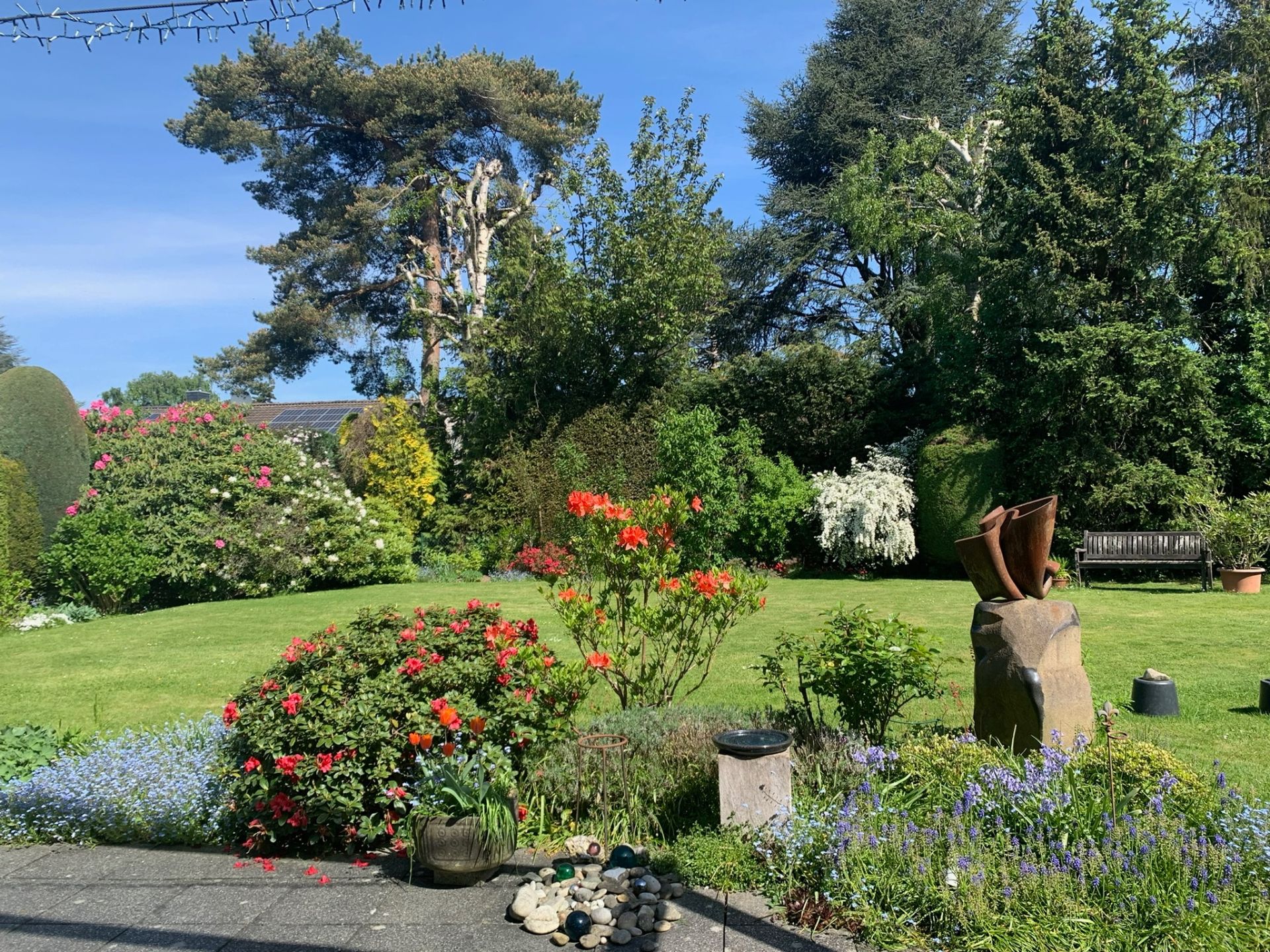 Grüner Garten mit Büschen, Bäumen, Blumen, Skulptur und einer Holzbank.