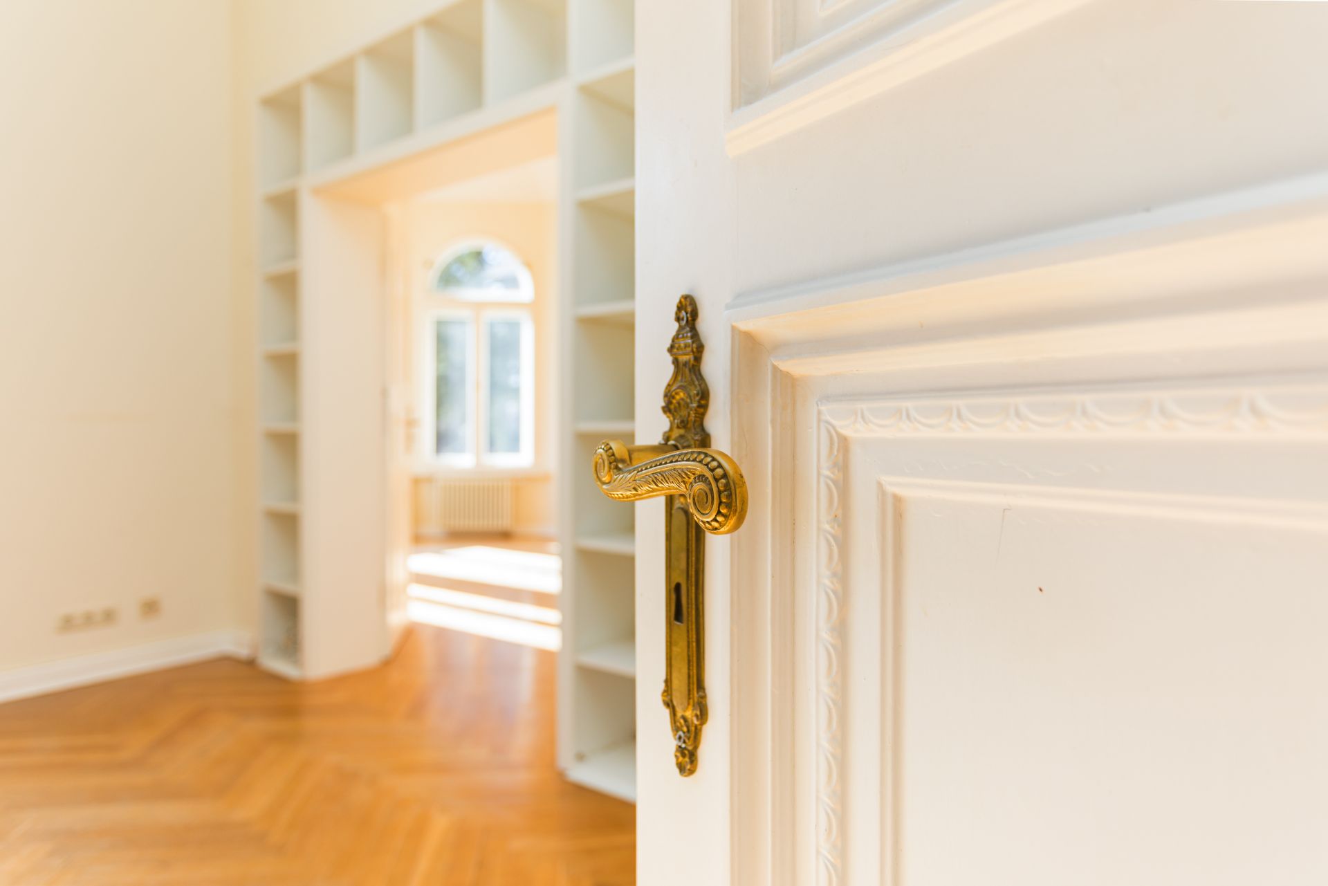 Offene weiße Tür mit goldener Klinke, Blick auf helles Zimmer mit Bücherregal und Parkettboden.