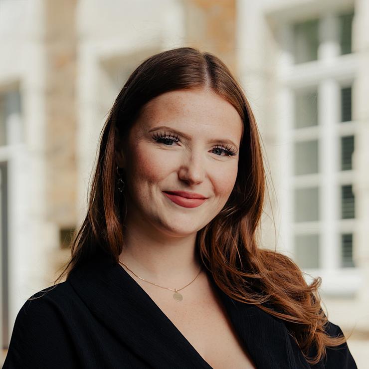 Eine junge Frau mit langen braunen Haaren steht vor einem Fenster im Hintergrund.