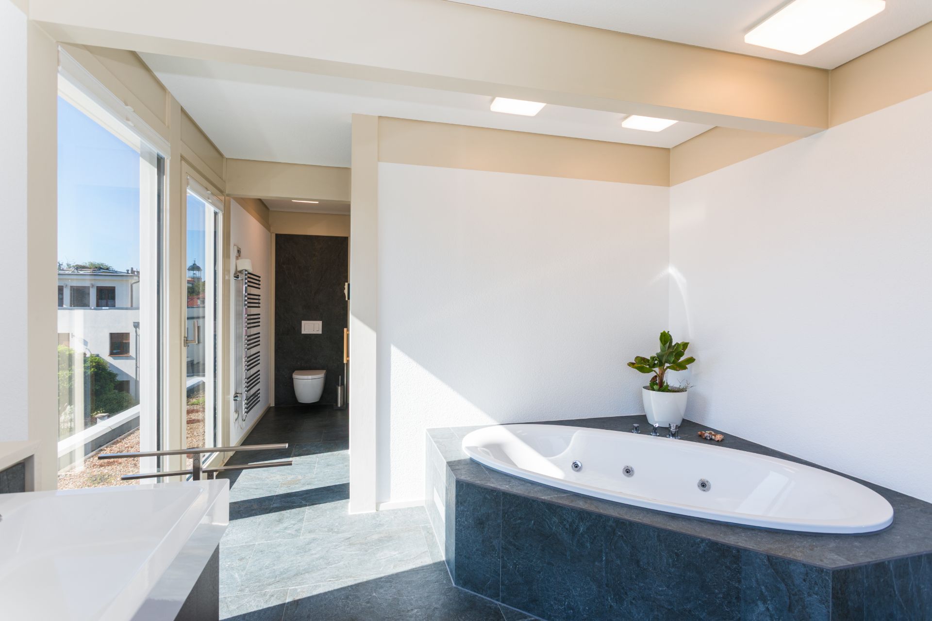 Helles Badezimmer mit großer Eckbadewanne, Pflanze und bodentiefen Fenstern.
