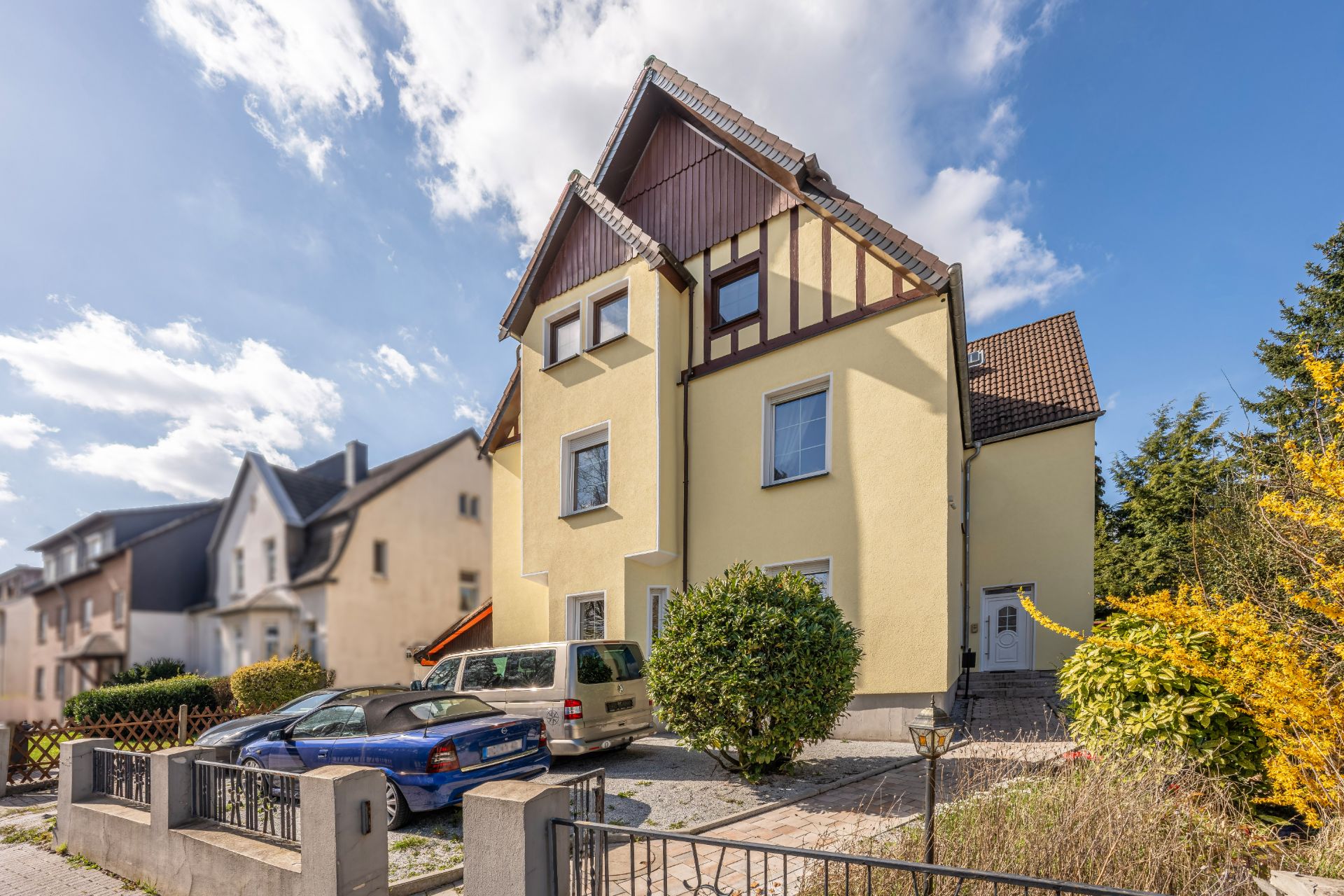 Wohnhaus mit gelber Fassade, zwei parkenden Autos und Sträuchern im Vorgarten.
