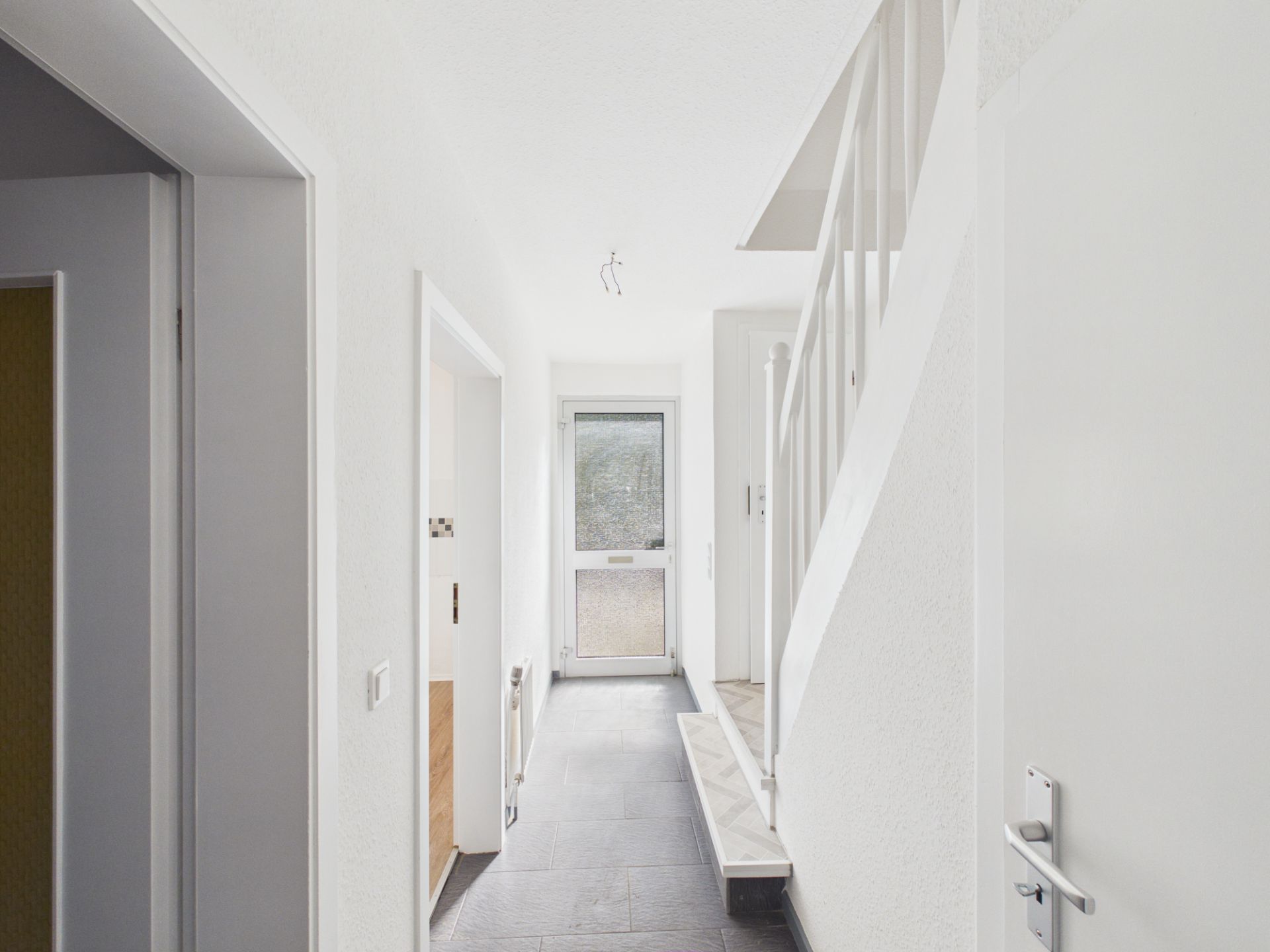 Heller Flur mit Treppe und Tür am Ende, weiße Wände und grauer Boden.