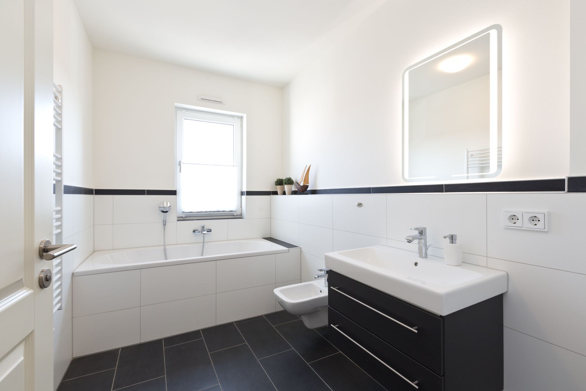 Modernes Badezimmer mit Badewanne, Waschbecken, beleuchtetem Spiegel und Fenster.
