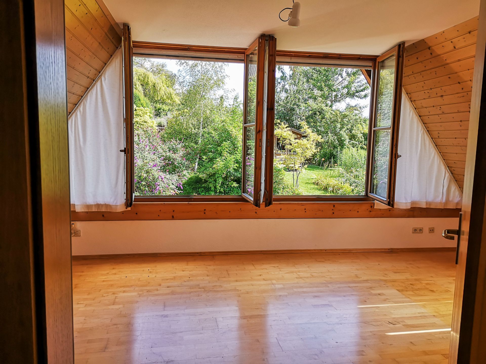 Heller Raum mit Holzfußboden und großen Fenstern, Blick auf grünen Garten.