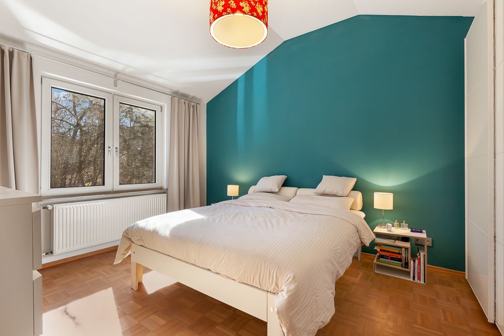 Helles Schlafzimmer mit Doppelbett, türkisfarbener Wand, großem Fenster und Nachttischen mit Lampen.