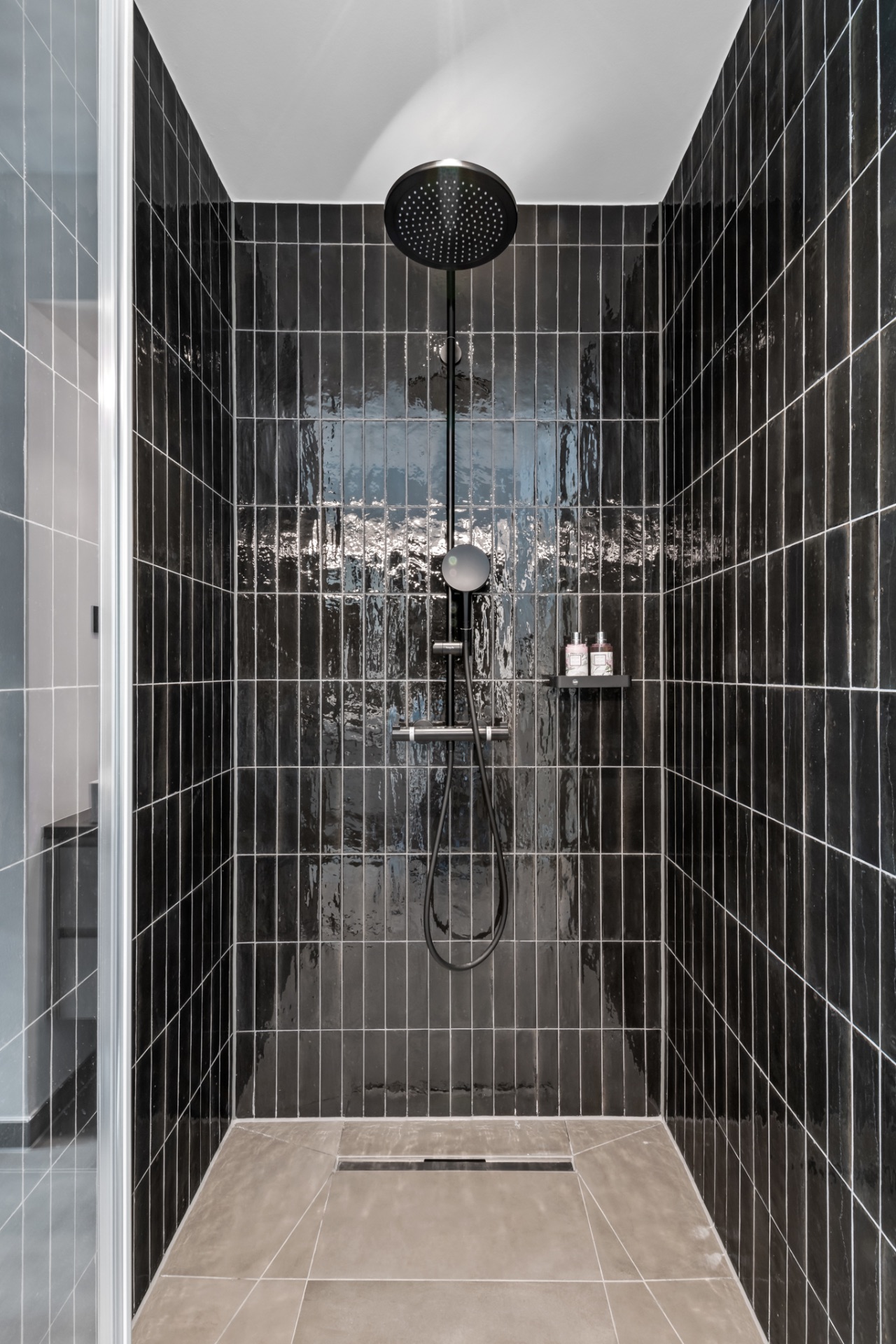 Moderne Dusche mit schwarzen Fliesen, großer Duschkopf und Ablage mit Duschprodukten.