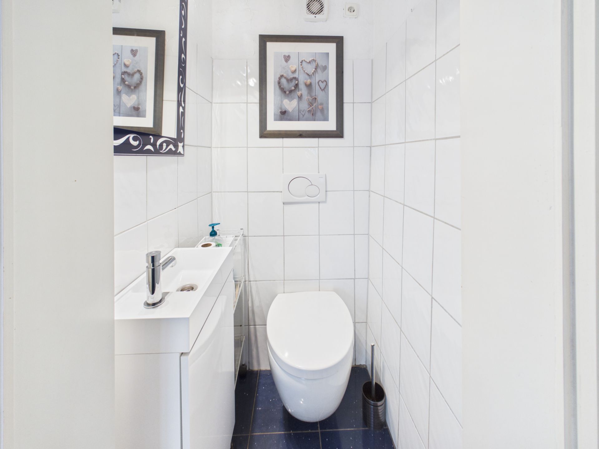 Kleines Badezimmer mit weiß gefliesten Wänden, WC, Waschbecken und Wandbild mit Herzmotiven.
