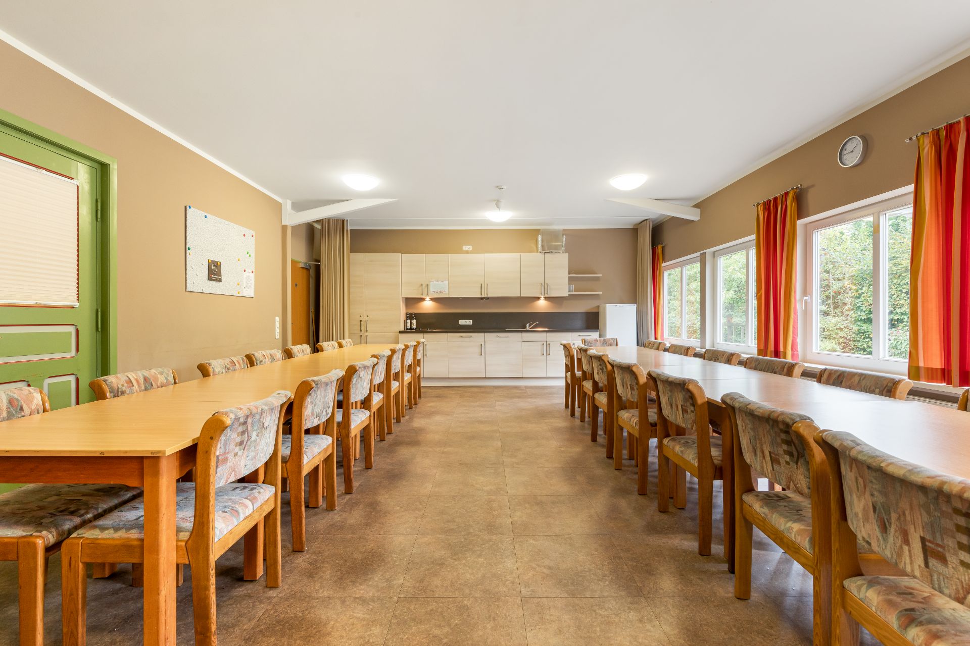 Großer Gemeinschaftsraum mit langen Holztischen, vielen Stühlen und offener Küche, Fenster mit roten Vorhängen.