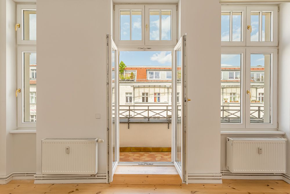 Helles Zimmer mit großen Fenstern und offener Tür zum Balkon, Blick auf Nachbargebäude.
