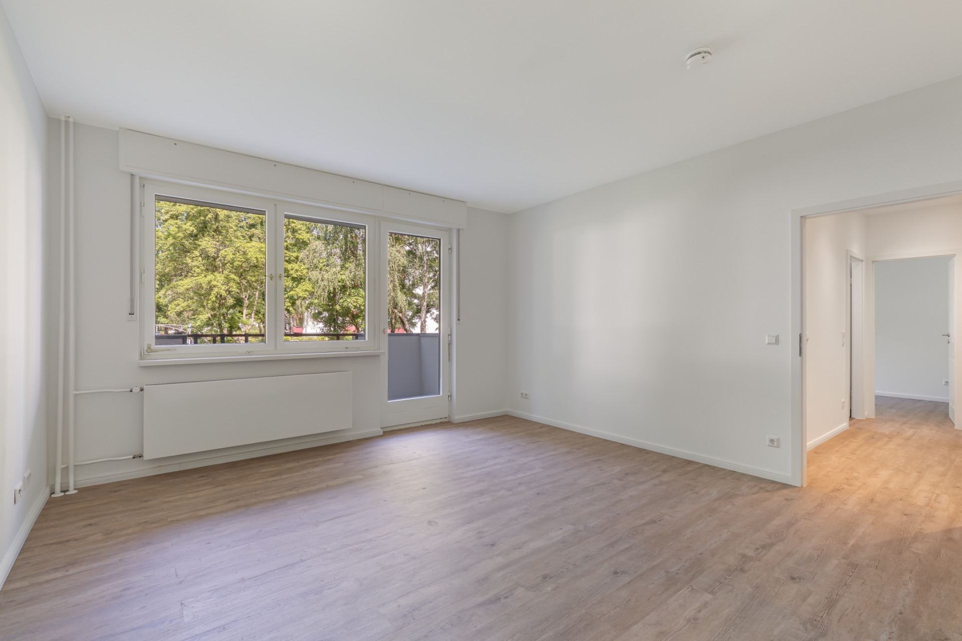 Leerer, heller Raum mit Holzfußboden, großen Fenstern und Zugang zu einem weiteren Zimmer.