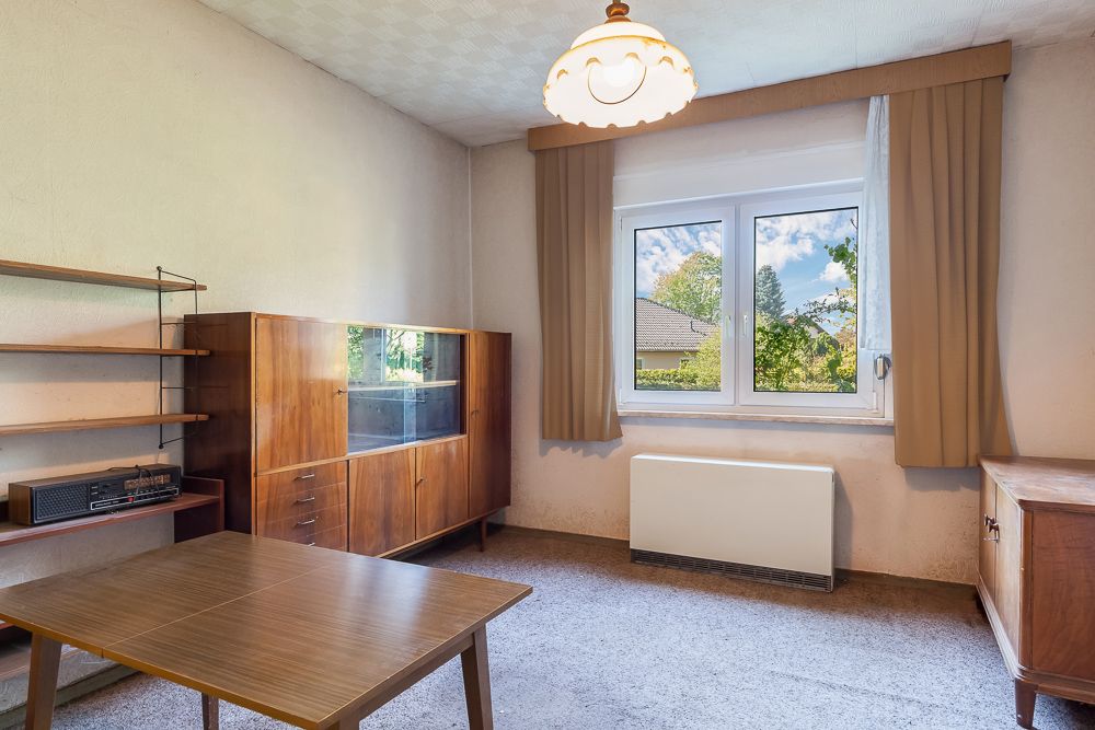 Wohnzimmer mit Holztisch, Sideboard, Regal, Teppichboden und Fenster mit Vorhängen.