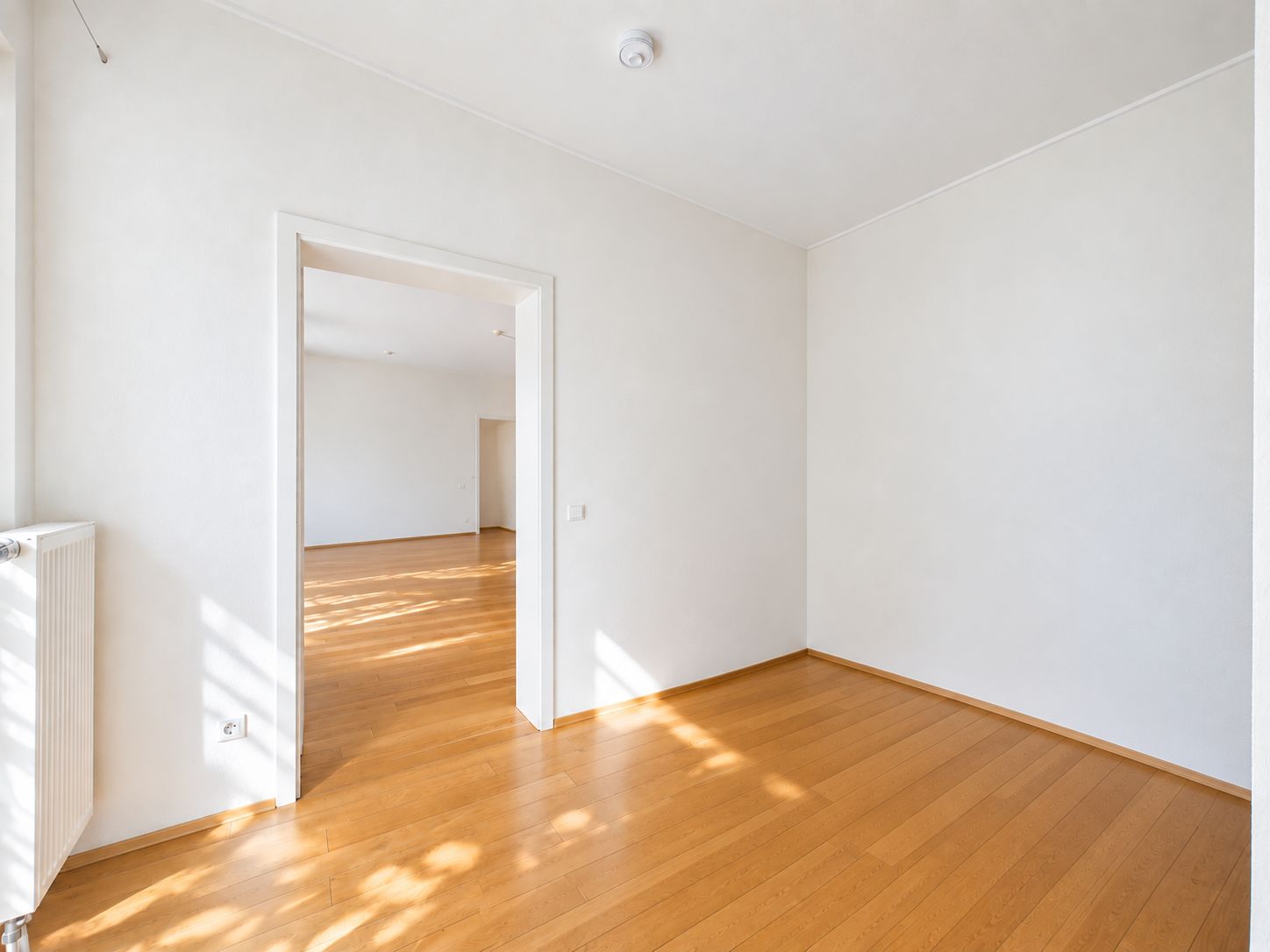 Heller Raum mit Holzfußboden und weißen Wänden, Türöffnung zu weiterem Zimmer.