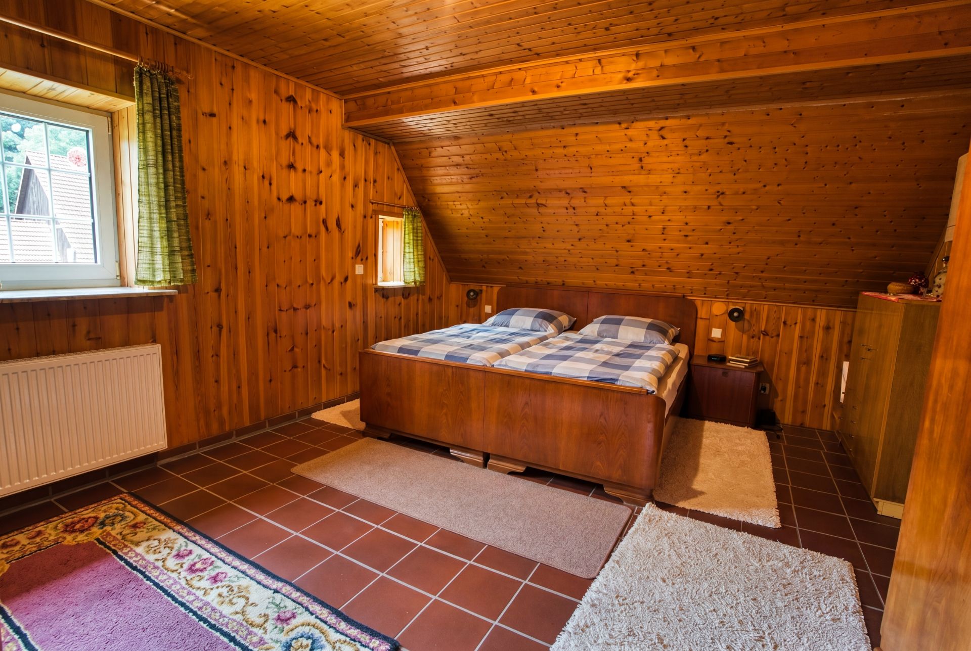 Schlafzimmer mit Holzdecke und -wänden, Doppelbett, Fenster, Teppichen und Fliesenboden.