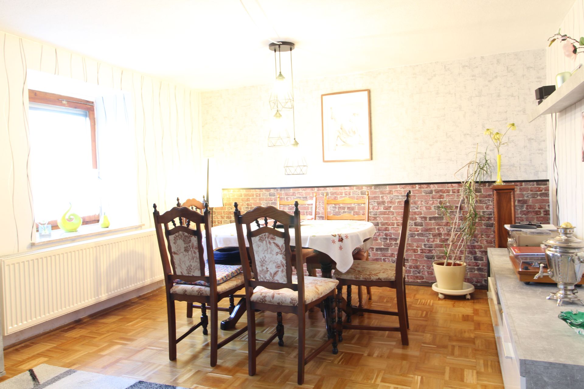 Esszimmer mit Holzboden, rundem Tisch und vier Stühlen, Pflanze in der Ecke, Fenster links.