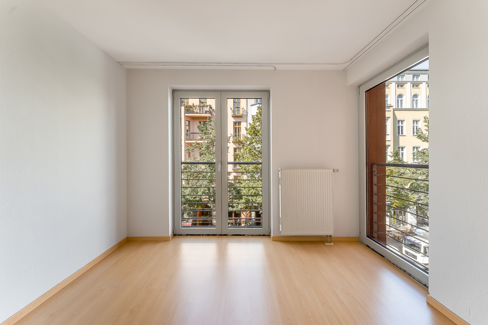 Heller, leerer Raum mit Parkettboden, großen Fenstern und Blick auf Wohngebäude.